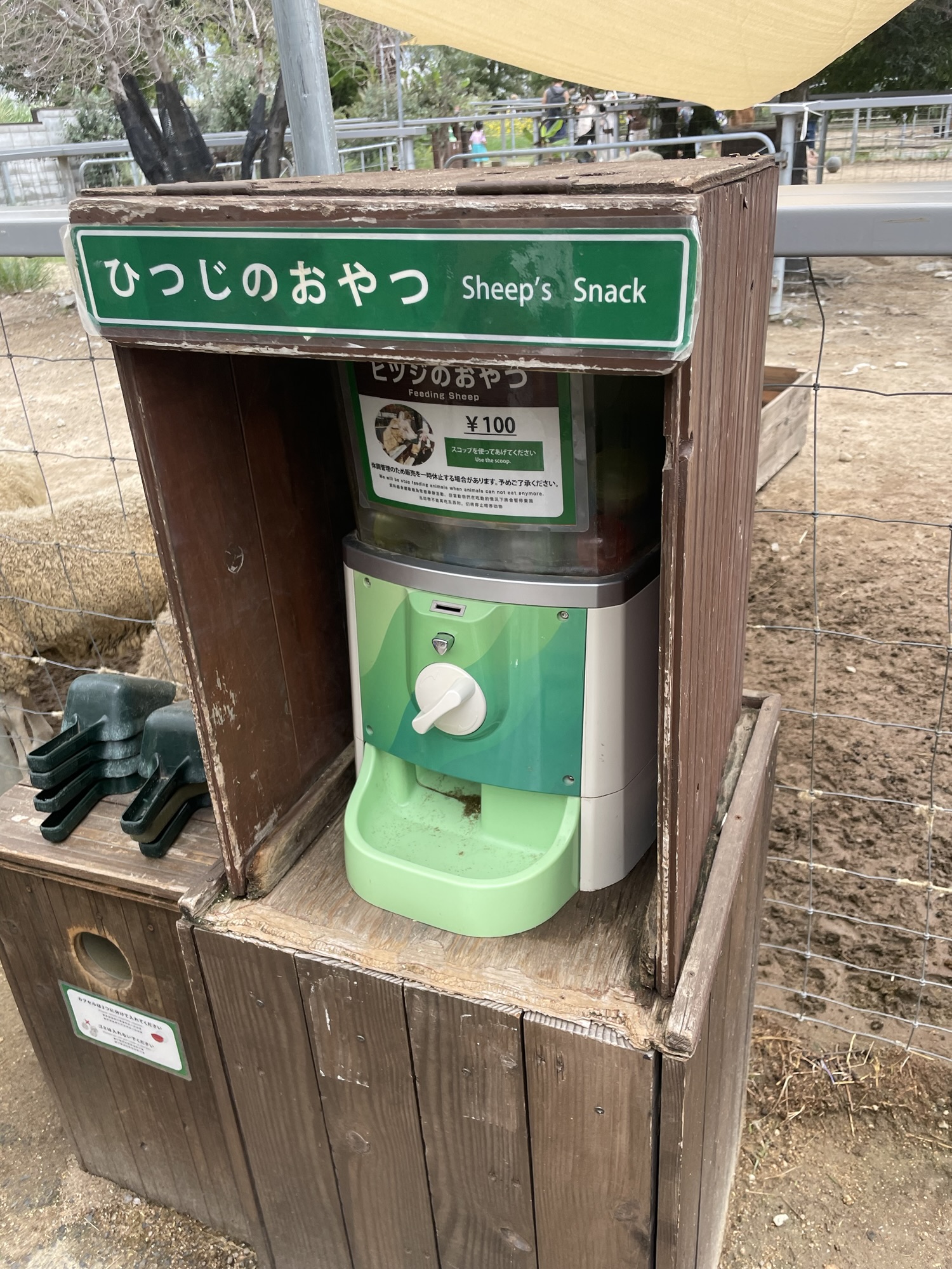 神戸どうぶつ王国の羊の餌販売機。子どもでも簡単に購入できるコイン式の自動販売機で、餌を手に取る様子が見える。